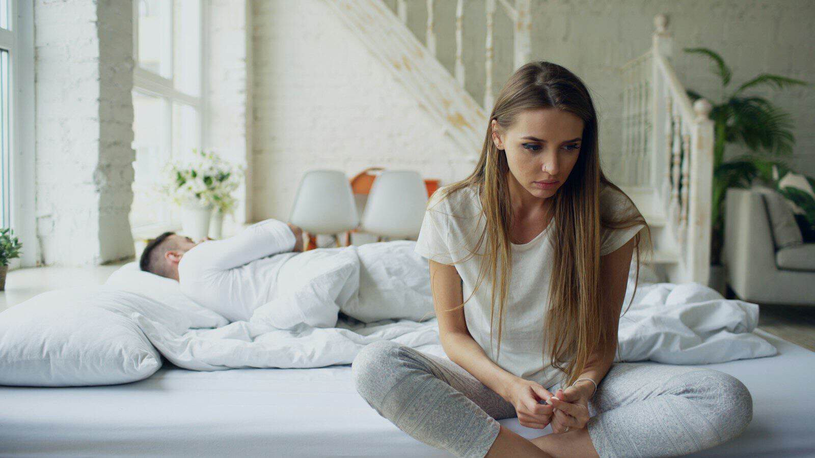 Woman sitting on bed, man sleeping behind her